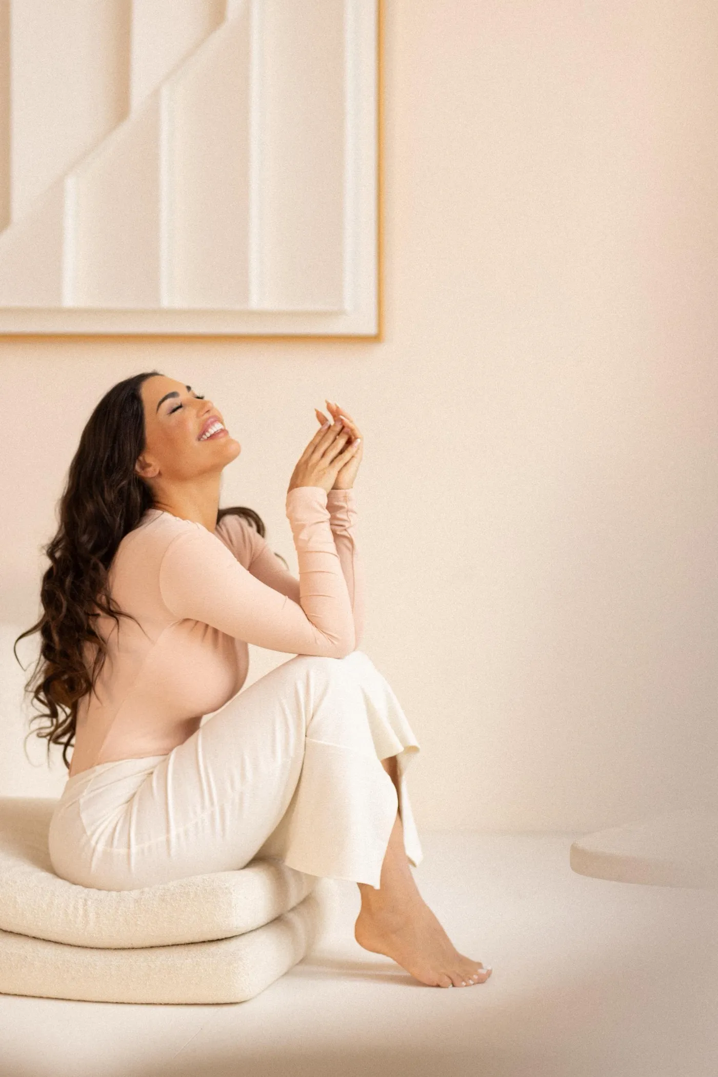 A woman in cream sits on the floor smiling, bathed in warm light