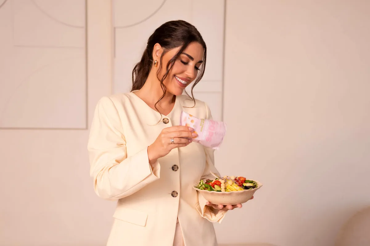 A smiling woman in a cream blazer holding a bowl of fresh food and a pink drink
